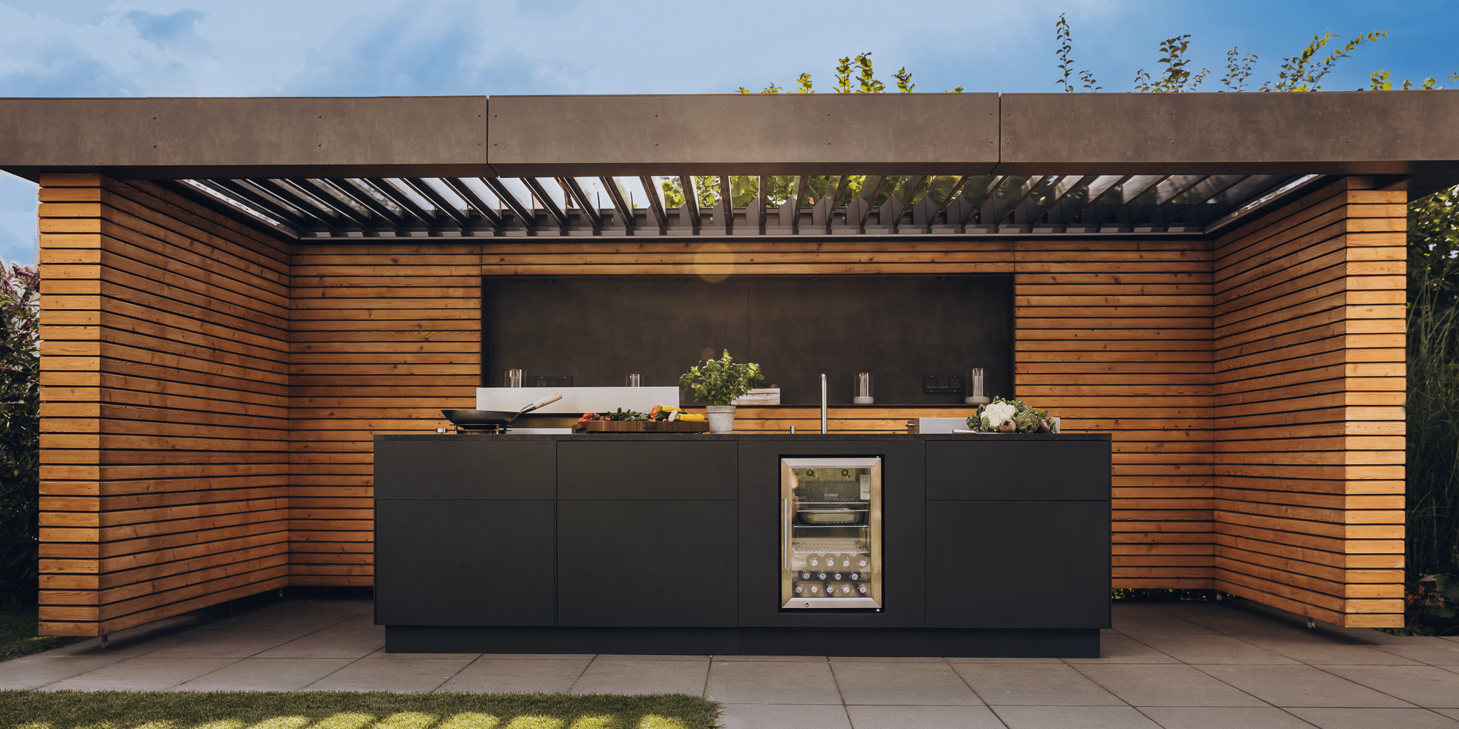 Outdoor kitchen with fire module, worktop and refrigerator surrounded by greenery
