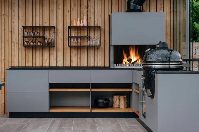 L-shaped outdoor kitchen with fire module, kamado grill and firewood compartments against timber cladding
