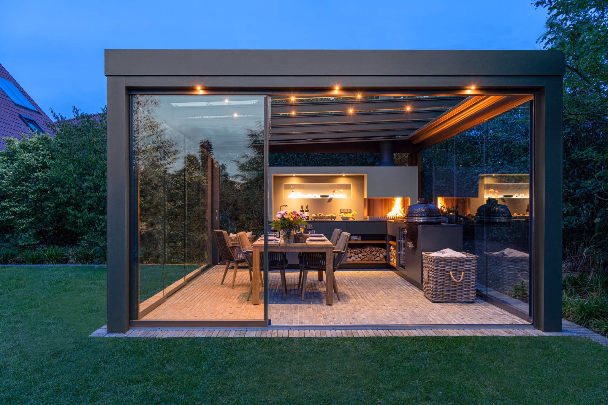 Outdoor kitchen with set dining table in glazed garden pavilion at dusk