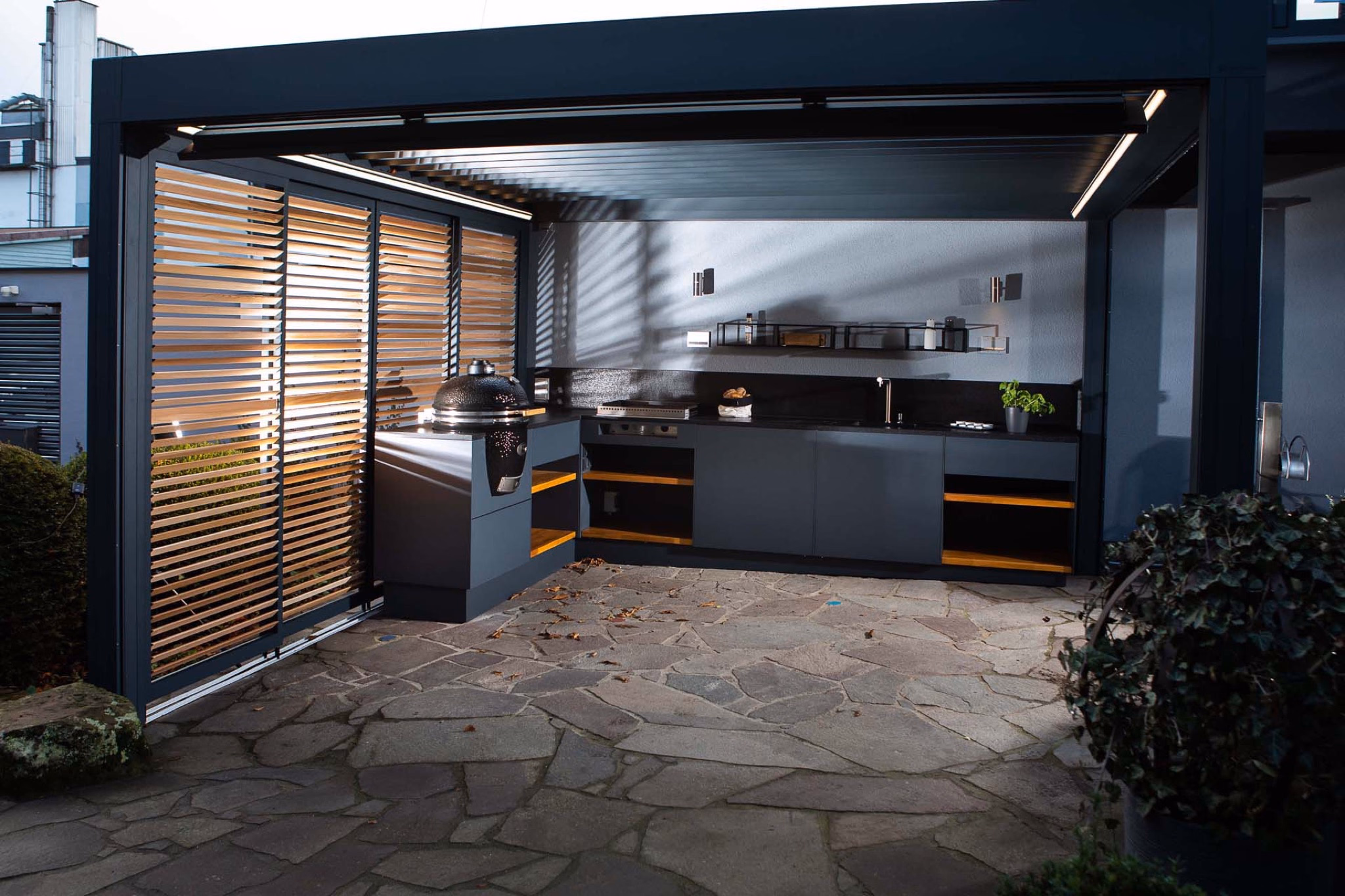 Outdoor kitchen under louvred roof with kamado grill on natural stone floor
